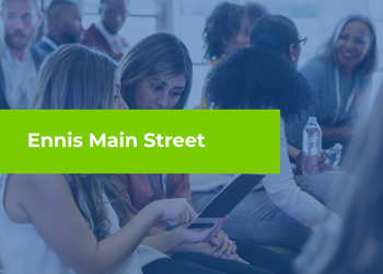 A group of people sitting together in a meeting or event setting, with a bright green banner across the center that reads “Ennis Main Street.”